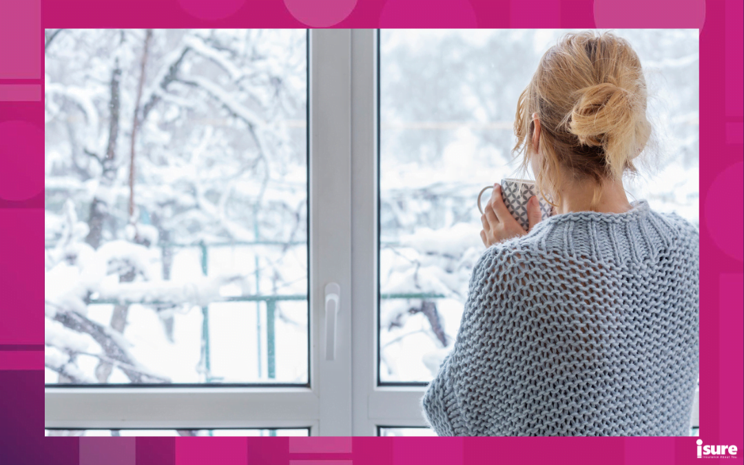 cut your heating bill - A woman in a cozy sweater stands at the picture window with a cup of drink in her hands