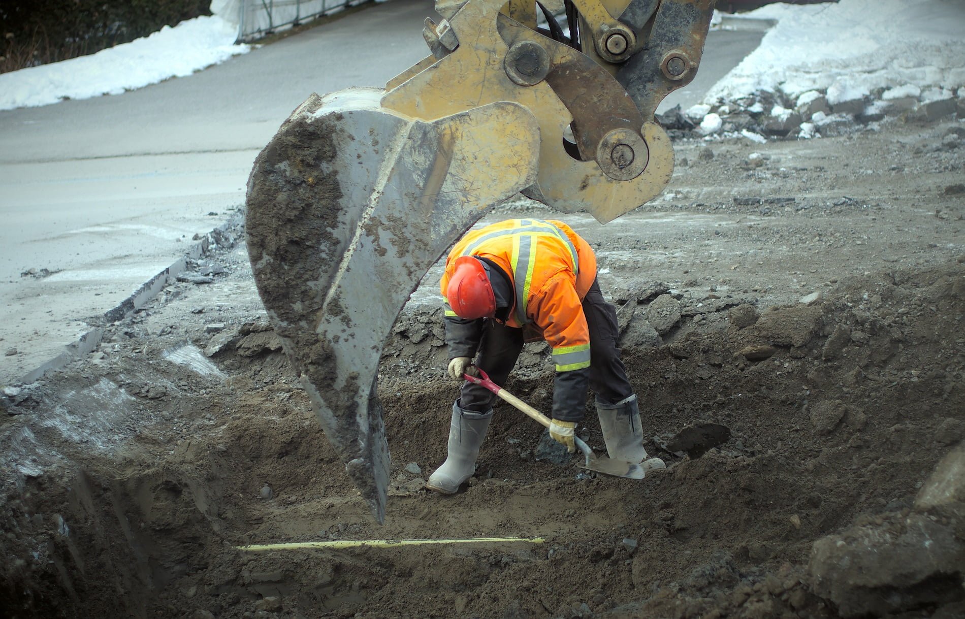 worker digging a hole with a shovel alongside an excavator inside the hole 