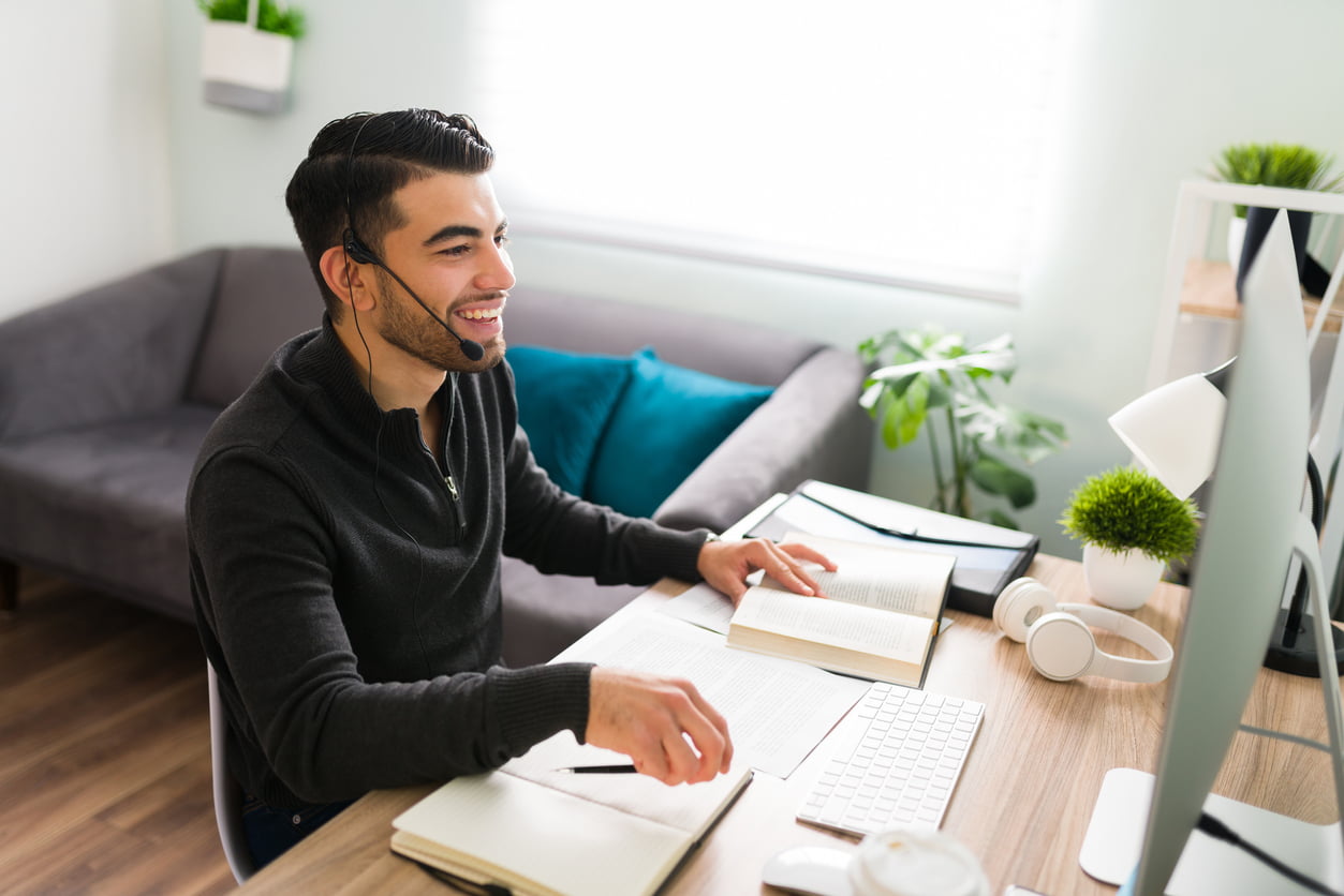 Good-looking young man sitting at his desk and wearing a headset. Smiling guy working as a translator or interpreter with a notebook and books - remote employees