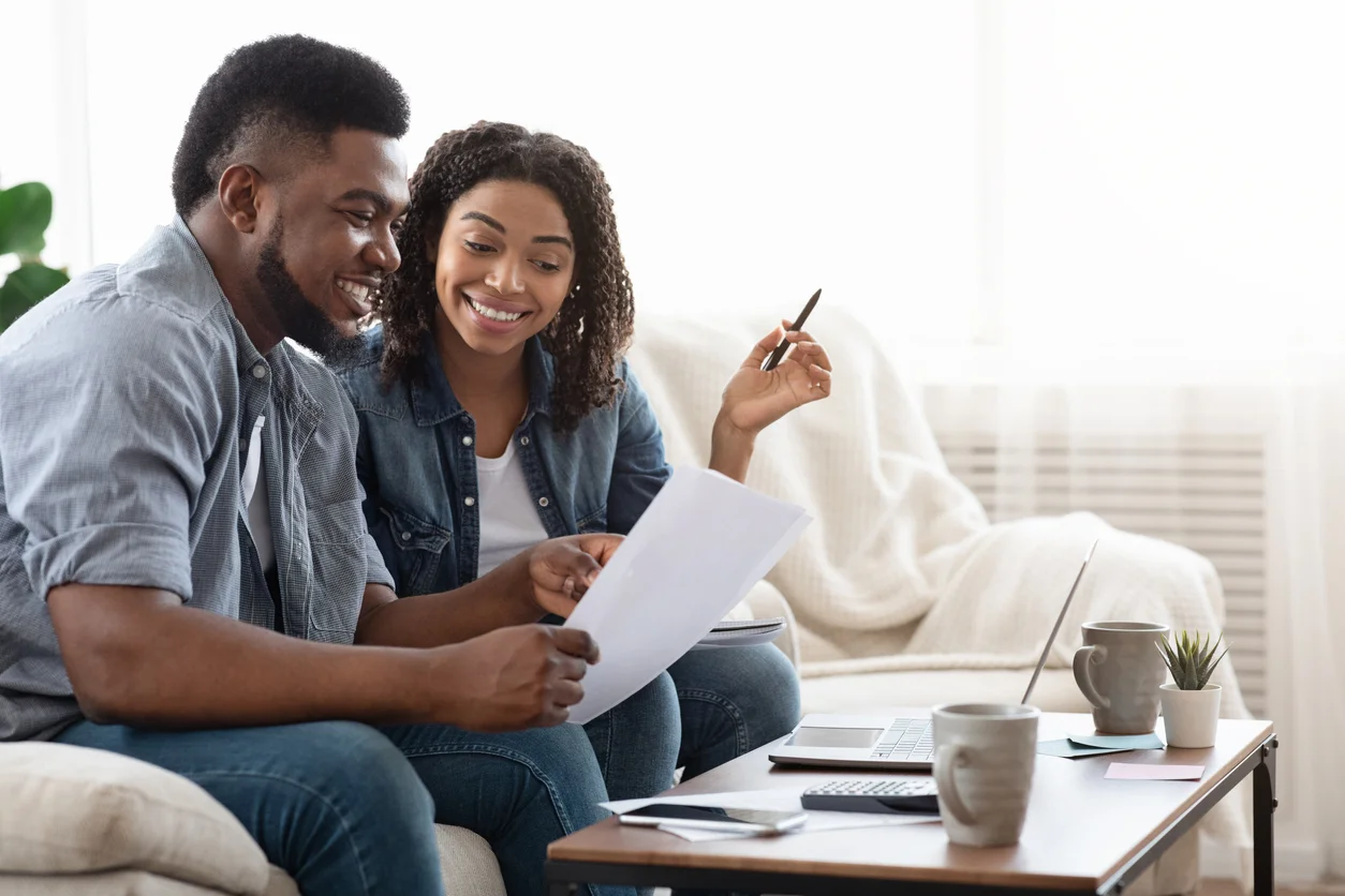 Young african american spouses checking documentation at home, reading insurance agreement or property certificate, sitting on couch together, smiling - home insurance questions for first-time home buyers