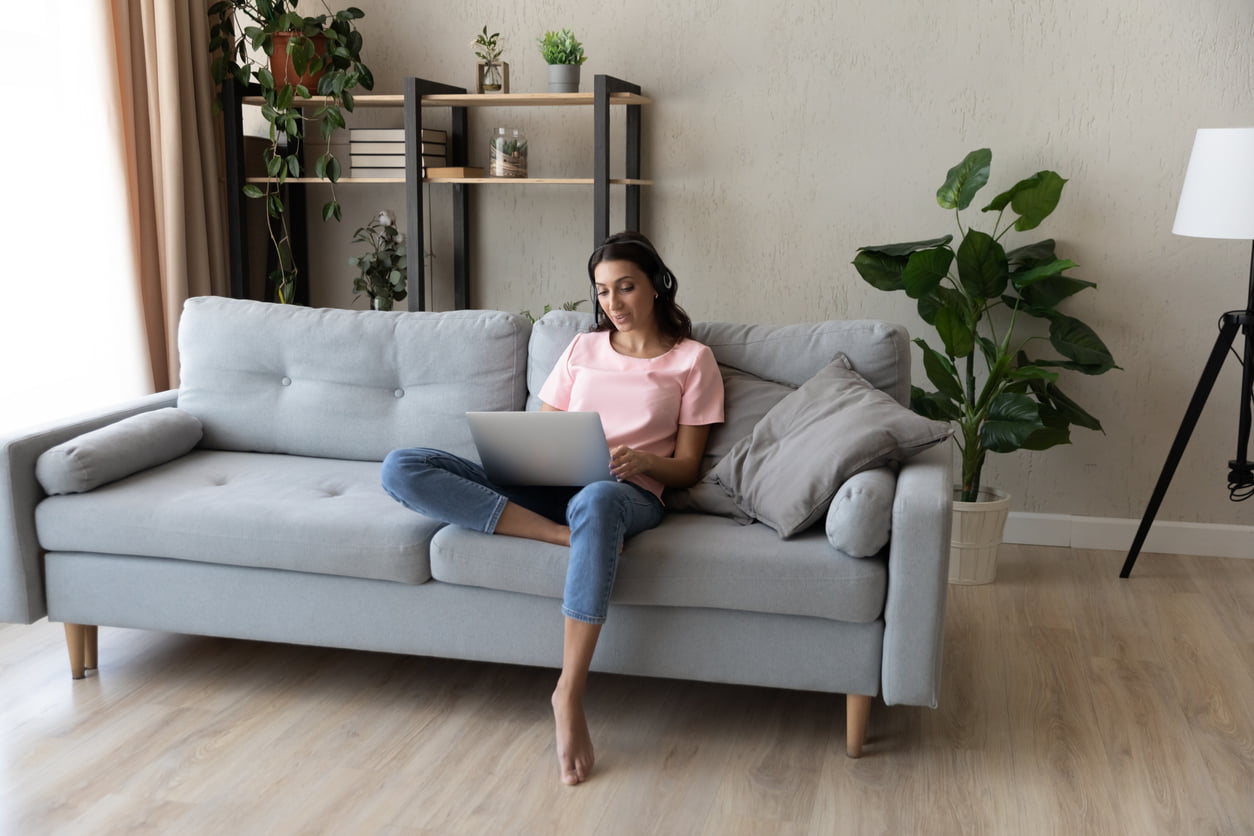 Full length focused young arabian indian woman wearing wireless headset with microphone sitting on sofa, holding distant online video call web camera meeting with colleagues, working from home. common myths about tenant insurance