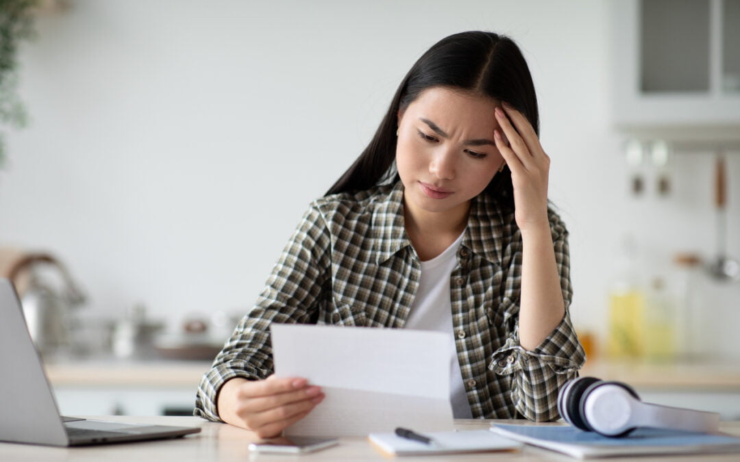 Can your insurer cancel your policy? Upset young asian woman sitting at table with modern laptop in kitchen, reading letter and touching her forehead, copy space. Sad lady holding document or bills, got bad news, home interior