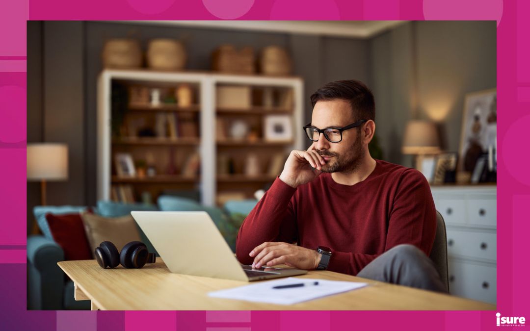 A busy, focused young adult male who works from home sitting at a desk and resting his head on his hand while working on a project on his laptop, focusing on his wellness.