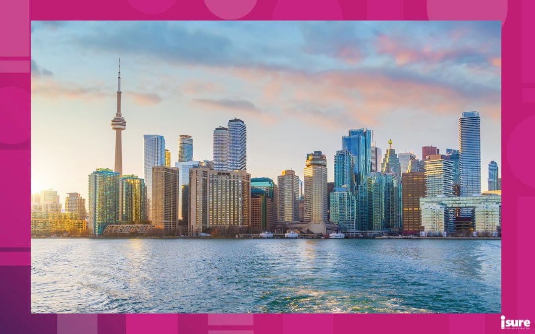 A wide-angle photo of the Toronto skyline across the water during sunset. The CN Tower stands prominently on the left among numerous skyscrapers.