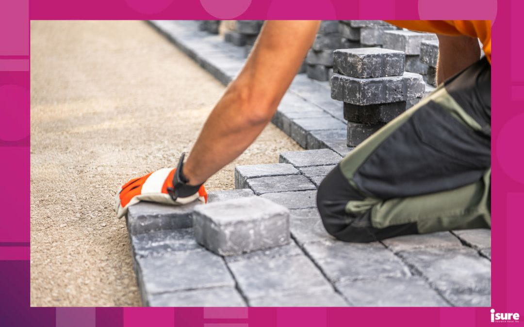 A hardscaping worker carefully positions paving stones on a pathway, focusing on alignment and precision. Wearing gloves, the individual kneels on a gravel surface, surrounded by stacks of stone blocks. The bright daylight highlights the texture of the stones and the surrounding area.