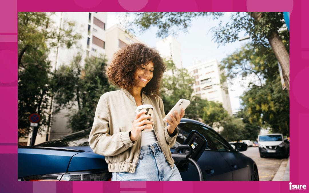 A woman stands outside her car, holding her smartphone and a coffee, smiling as she looks at her phone.